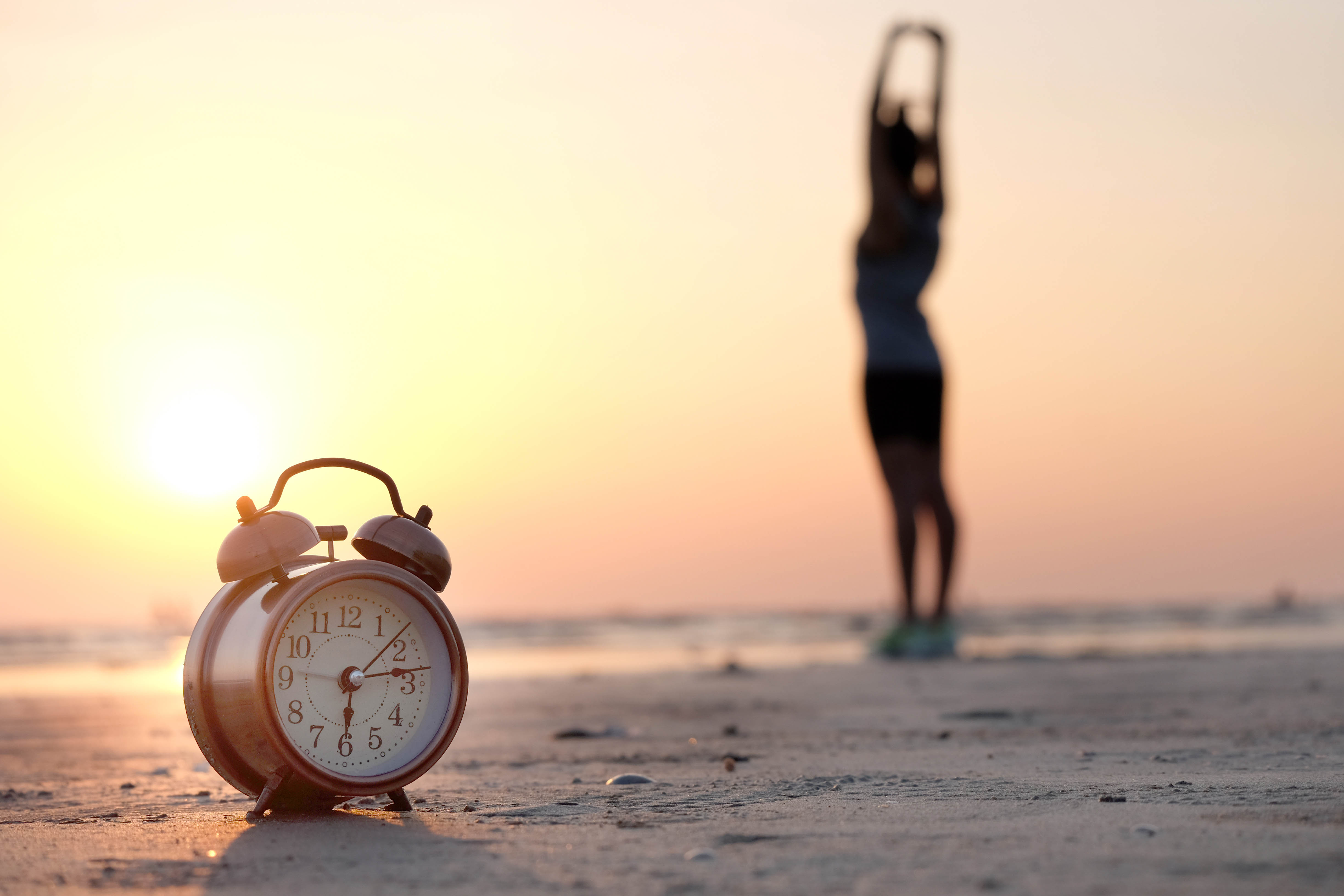 Woman stretching on the beach facing the sunrise, clock in the foreground showing that it is 6:15am